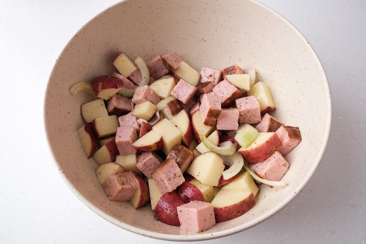 pieces of ham potatoes and onions covered in oil and spices in white mixing bowl.