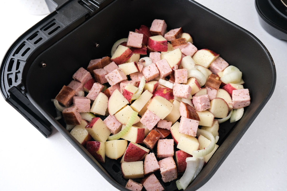 raw potato chunks and ham in black air fryer tray on white counter.