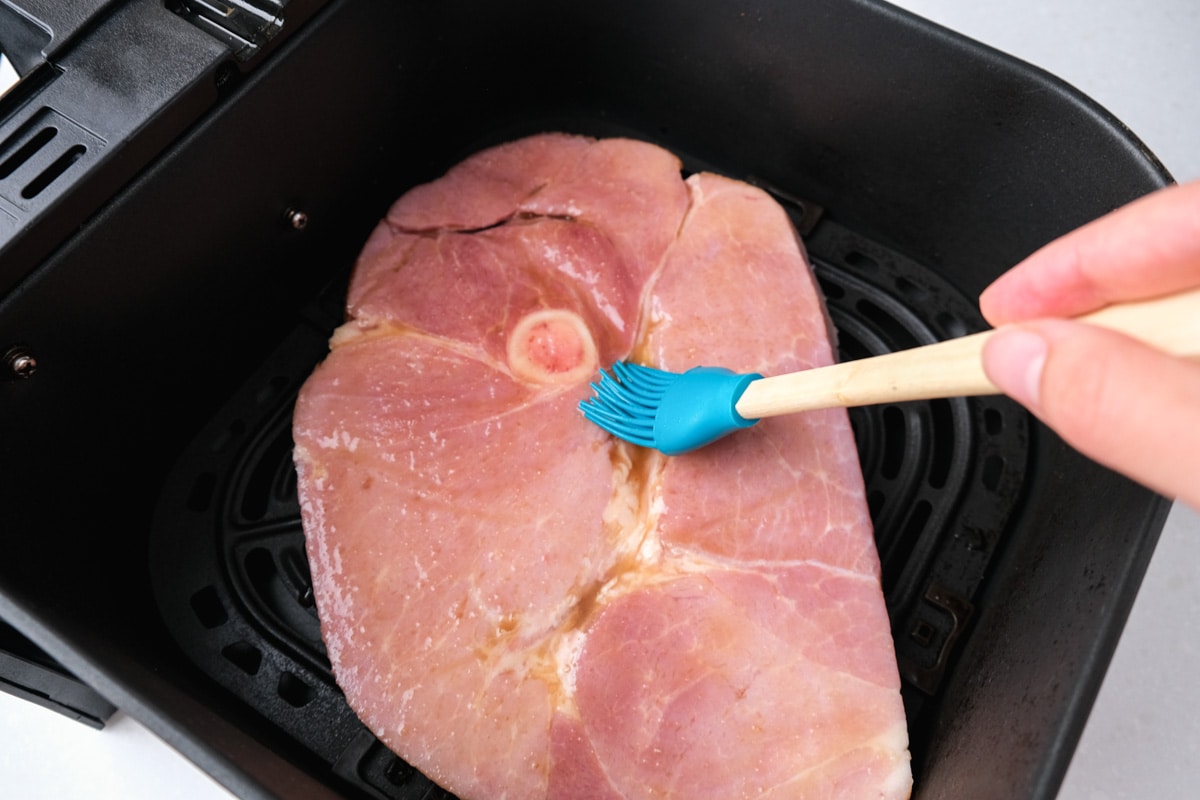 blue tipped marinade brush brushing glaze on ham steak sitting in black air fryer basket.