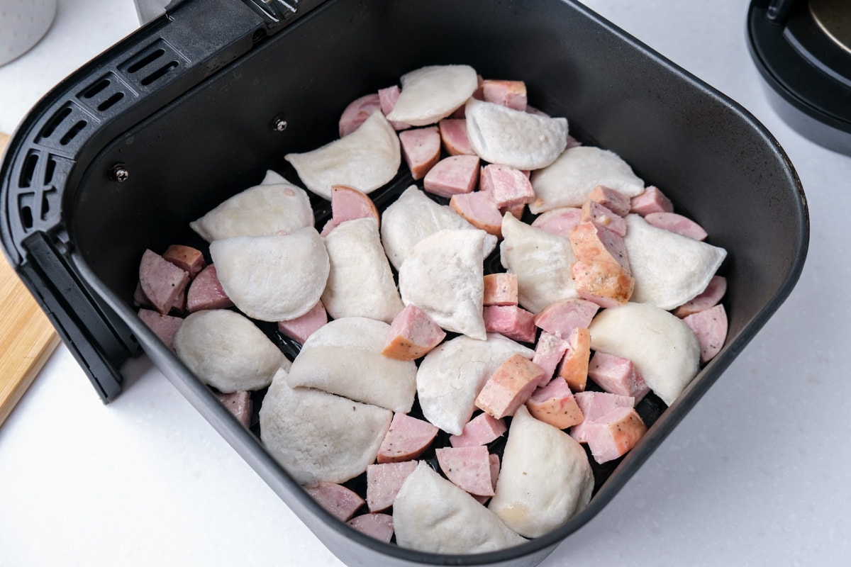 raw pierogies and kielbasa in black air fryer tray on white counter top.
