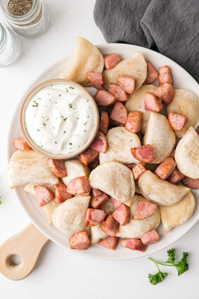pierogies and cooked kielbasa on plate with sour cream all sitting on wooden board with grey cloth beside.