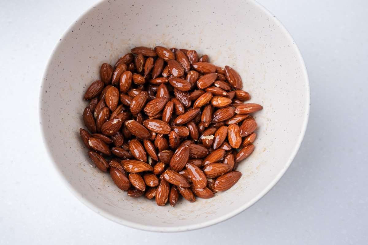 raw almonds in white mixing bowl covered in oil and salt sitting on white counter.