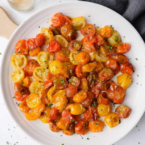 white plate of roasted cherry tomatoes with wooden board under and grey cloth beside.