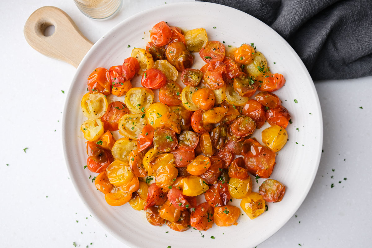white plate of roasted cherry tomatoes with wooden board under and grey cloth beside.