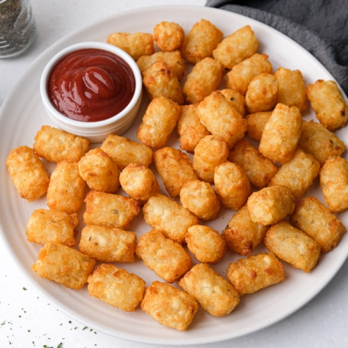 cooked tater tots on white plate on counter with ketchup beside in dipping bowl.