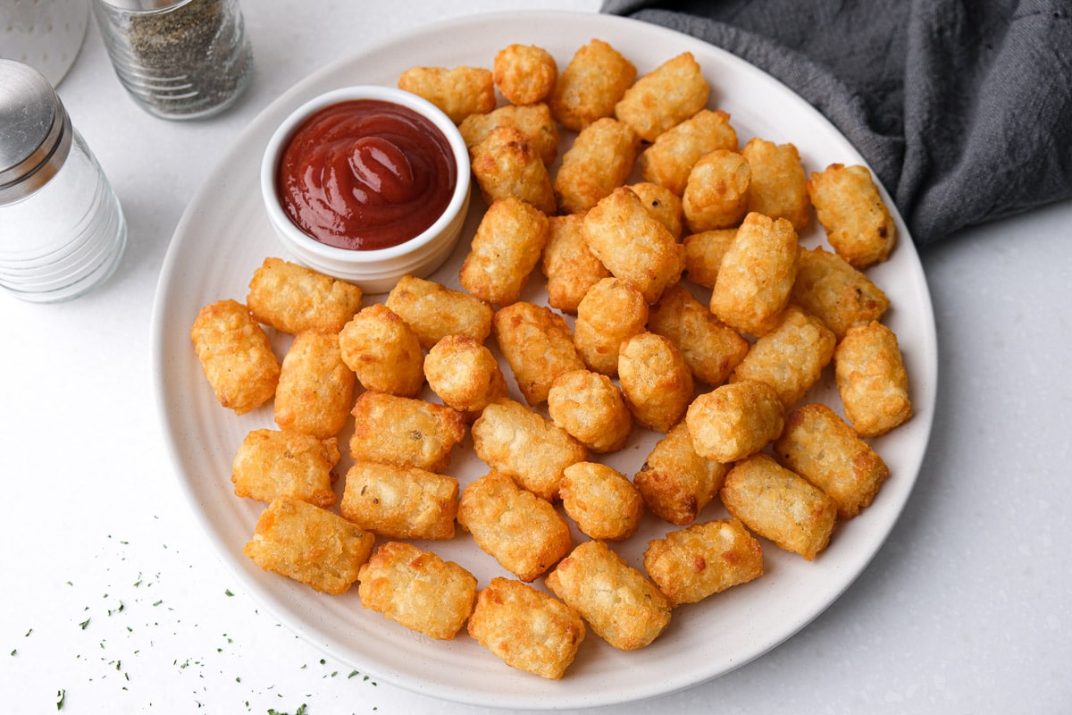 cooked tater tots on white plate on counter with ketchup beside in dipping bowl.