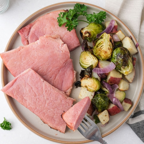 slices of ham on plate with vegetables beside on white counter top with dish towel.