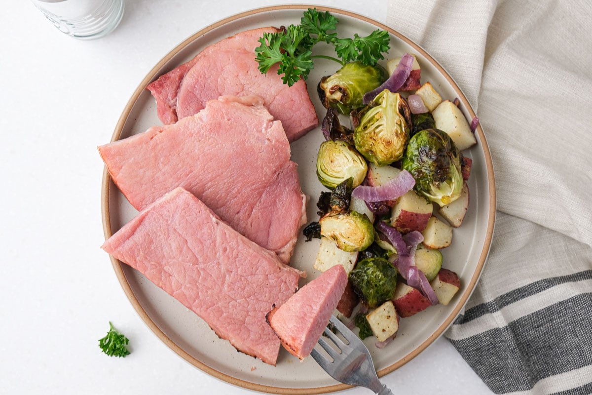 slices of ham on plate with vegetables beside on white counter top with dish towel.