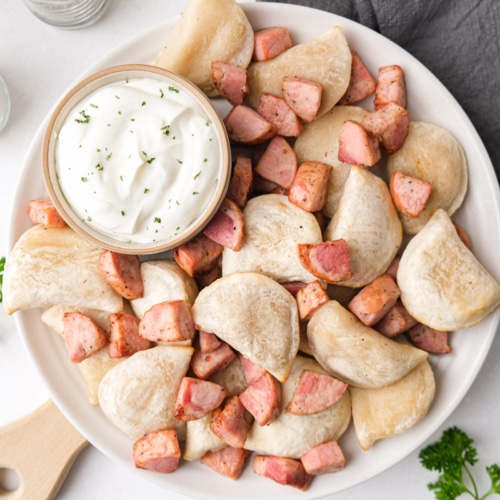 plate of cooked pierogies and kielbasa pieces with dish of sour cream beside.