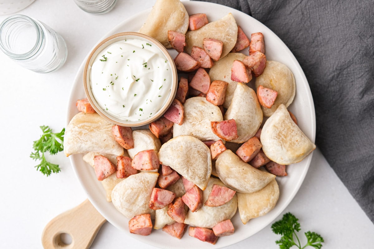 plate of cooked pierogies and kielbasa pieces with dish of sour cream beside.