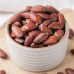 roasted almonds in small dish sitting on wooden board with other almonds around.