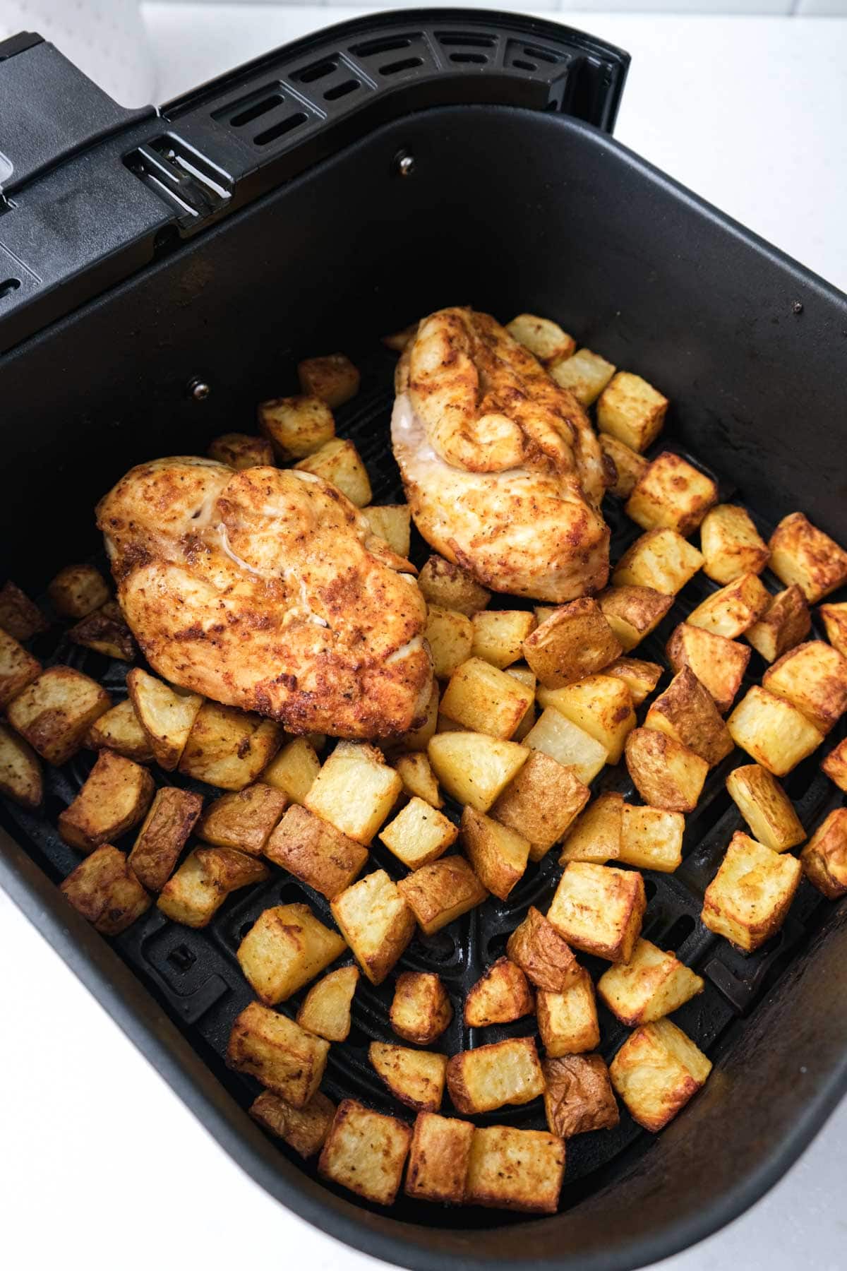 cooked chicken breast sitting on bed of cooked potatoes in black air fryer tray on white counter.
