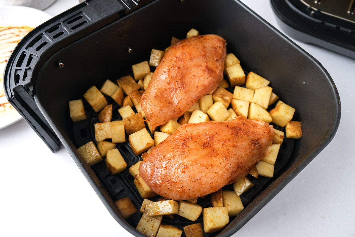 two raw chicken breast covered in spices sitting on bed of raw potatoes in black air fryer tray.
