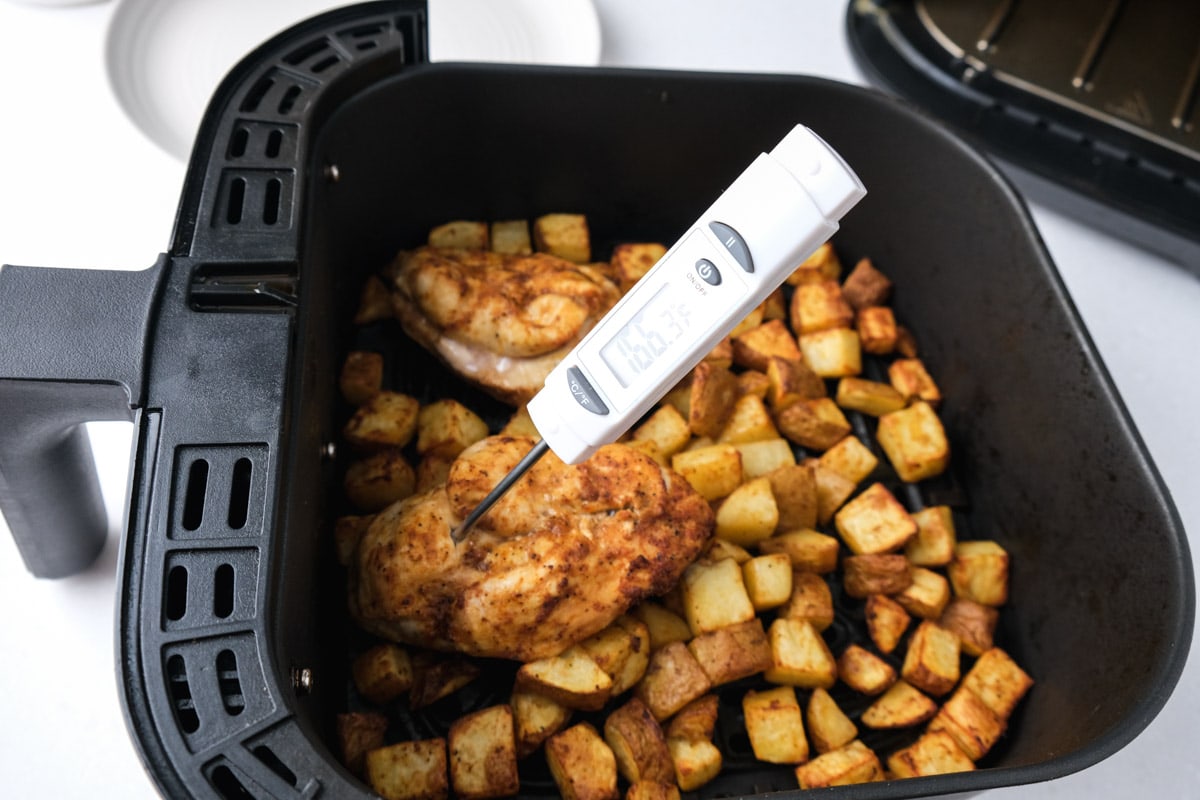 digital thermometer sticking out of cooked chicken breast in air fryer basket with potatoes around.