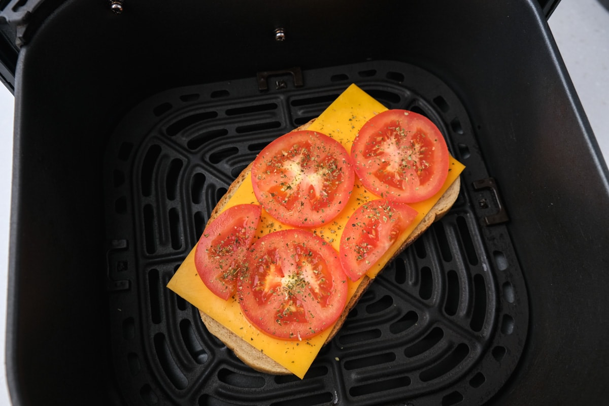 slice of bread sitting in black air fryer tray with layer of cheese slices and slices of tomato on top.