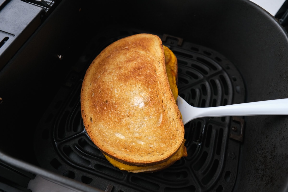 crispy sandwich sitting in black air fryer tray with white spatula undeneath.