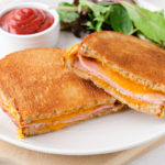 air fried grilled cheese and ham sandwich cut in half on white plate with ketchup and greens behind.