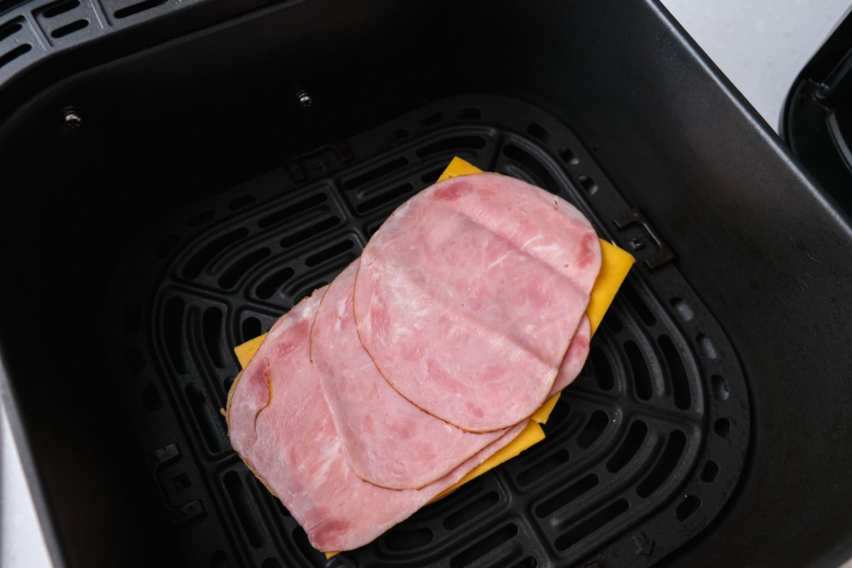 slices of ham and cheese sitting on open face bread in black air fryer tray.
