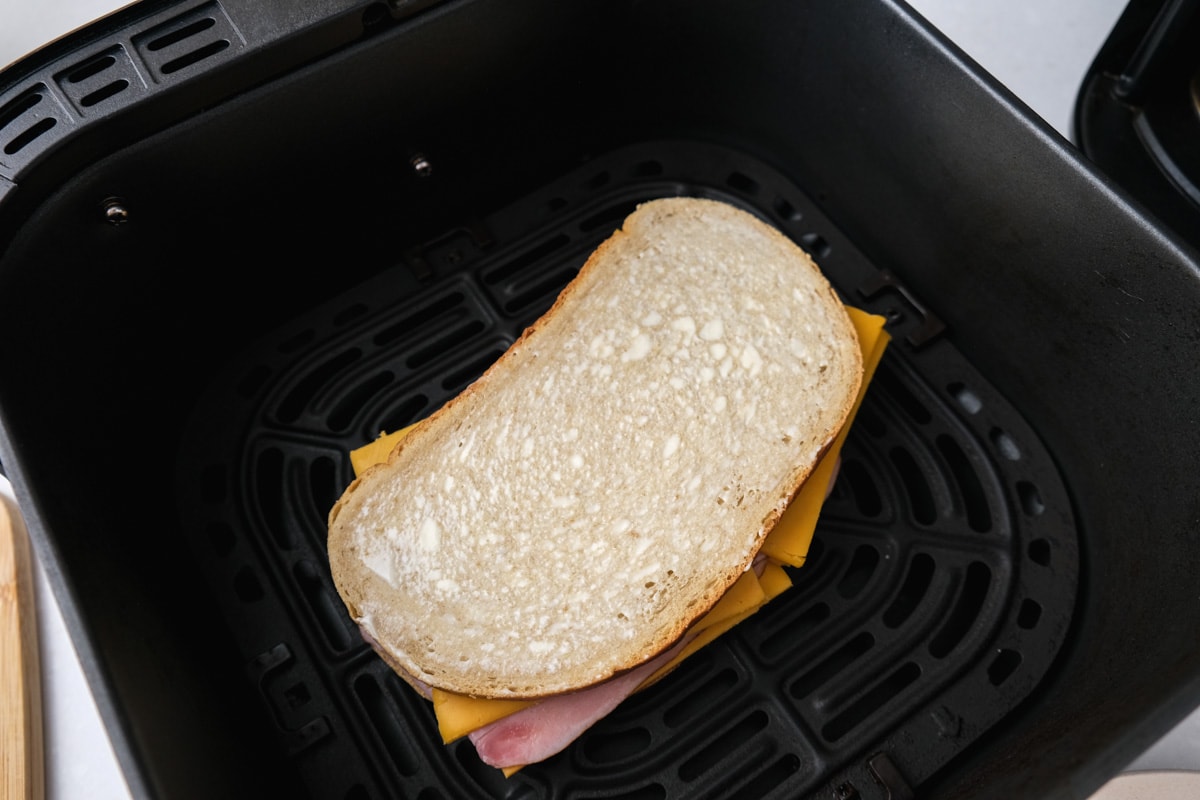 piece of rye bread with mayo on top sitting on top of slices of ham and cheese in black air fryer tray.