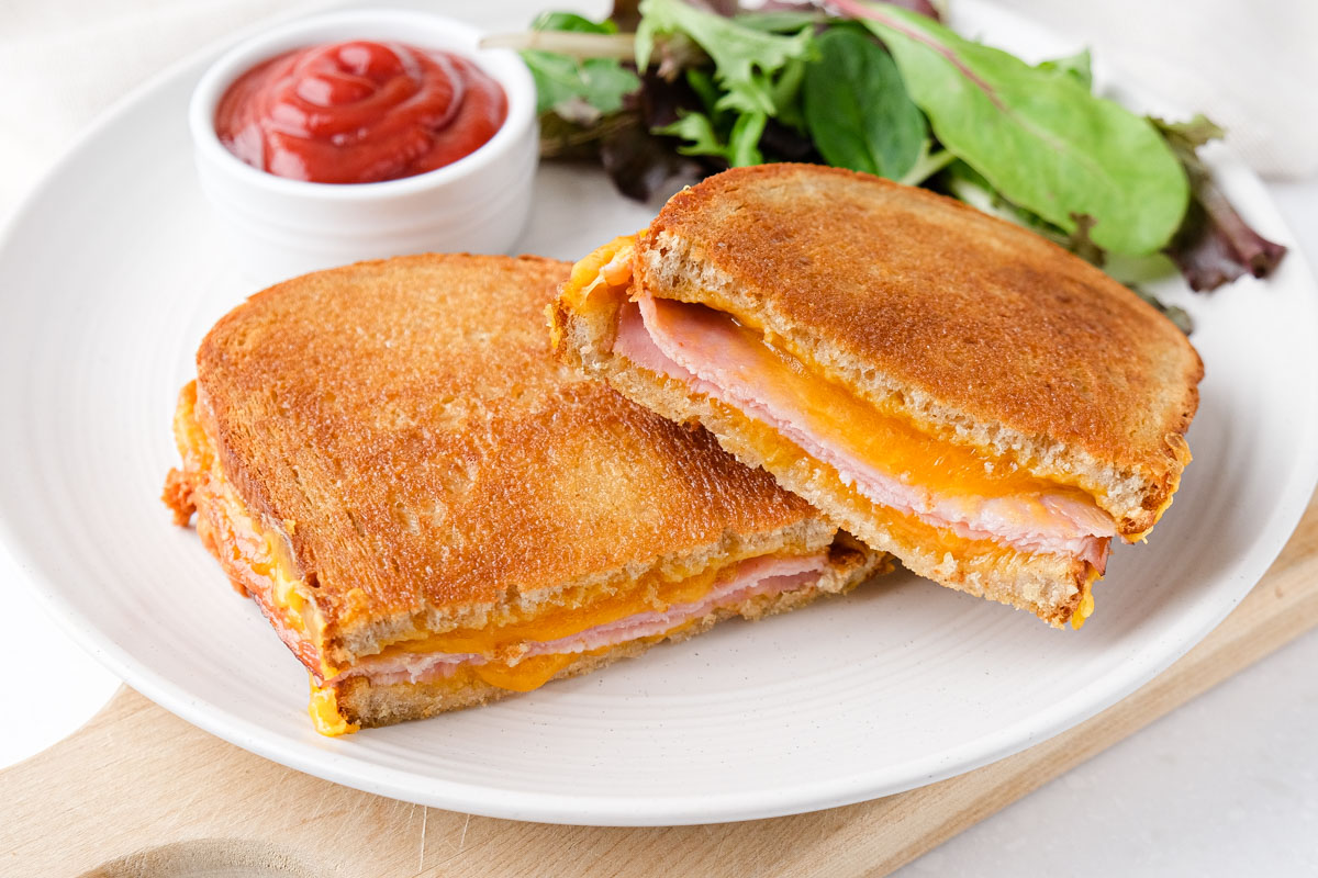 air fried grilled cheese and ham sandwich cut in half on white plate with ketchup and greens behind.