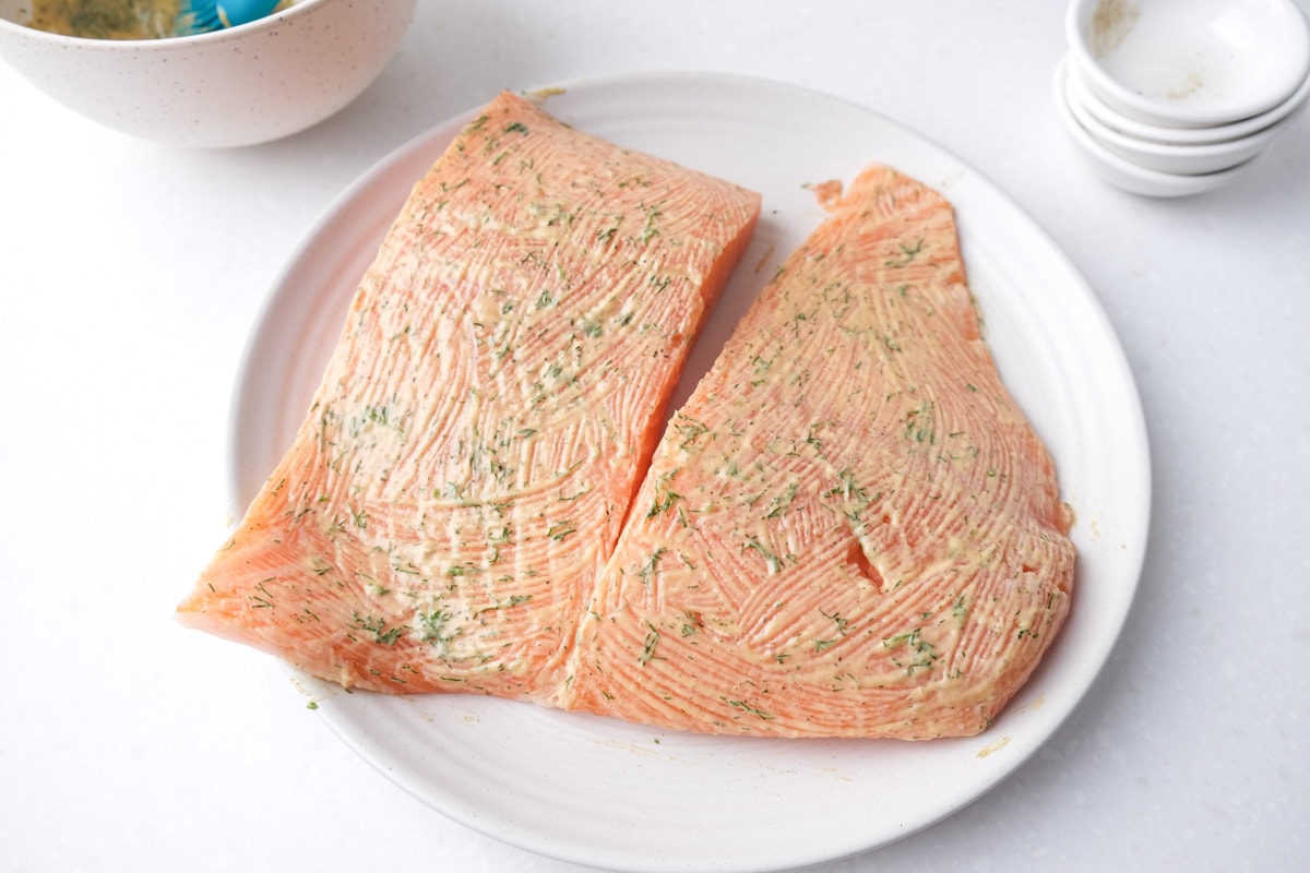raw salmon fillets sitting on white plate covered in mustard marinade with dill.