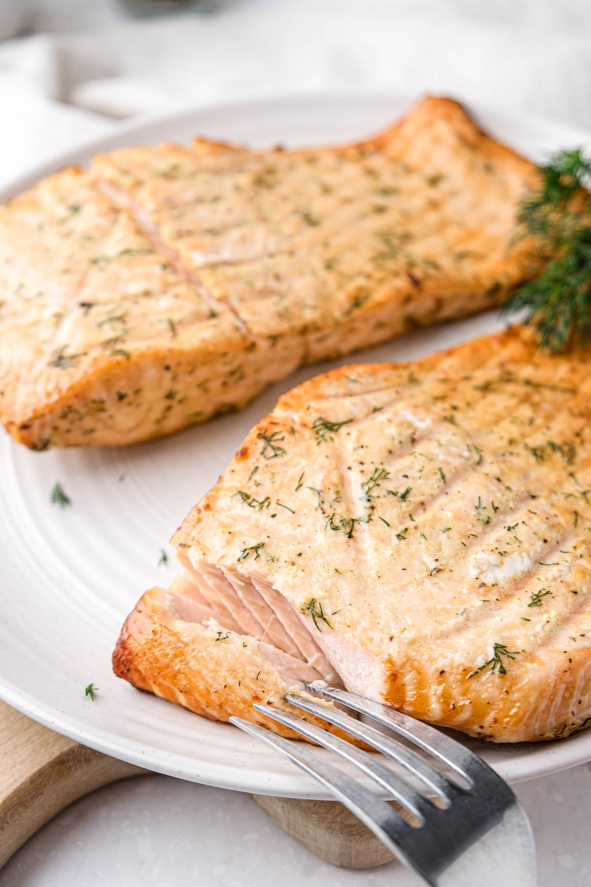 two fillets of cooked salmon with dill on top sitting on white plate with silver fork.