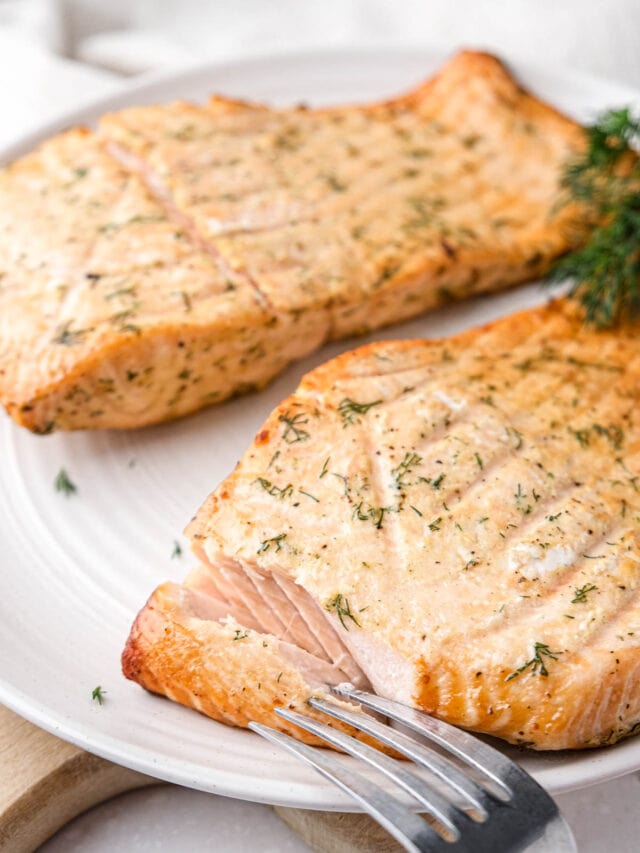 two fillets of cooked salmon with dill on top sitting on white plate with silver fork.