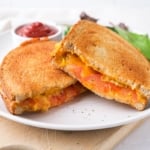 grilled cheese and tomato cut in half on white plate with ketchup and greens behind.