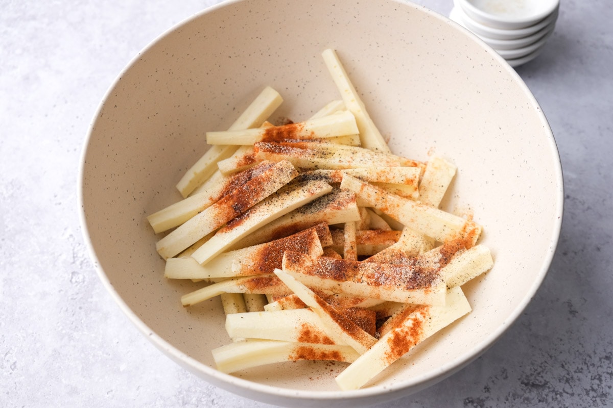 raw parsnip fries in white mixing bowl with spices and oil on top but not mixed in.