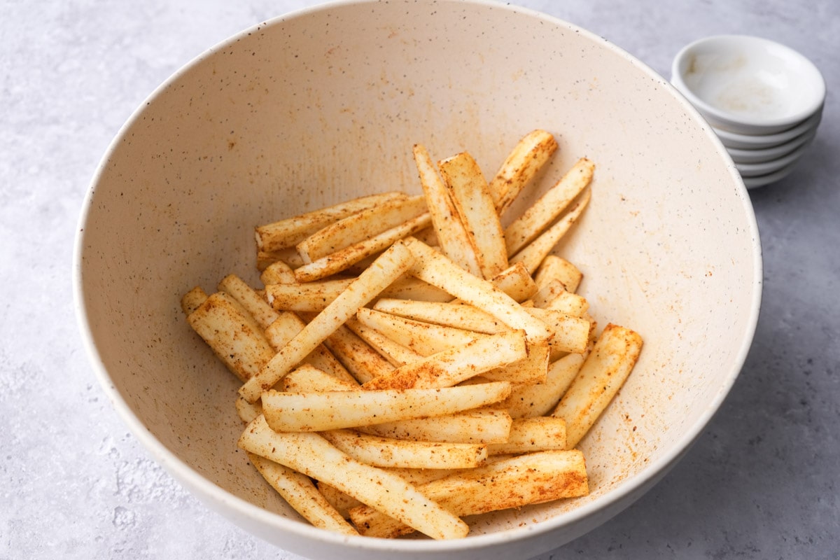 raw parsnip fries coated in spices in white mixing bowl.