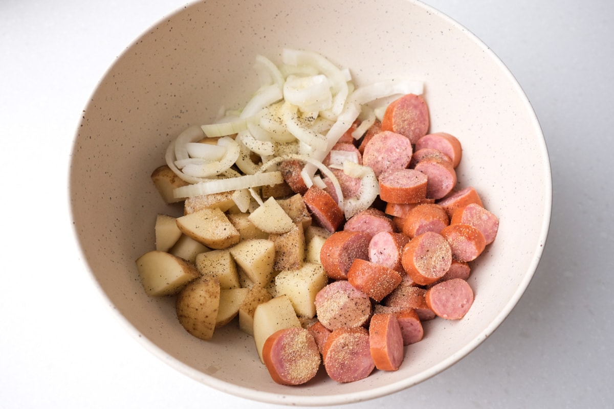 cut up onions potatoes and sausages in white mixing bowl covered in spices.