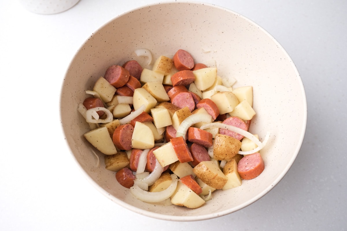 diced onions sausage and potatoes in white mixing bowl tossed in oil and spices.