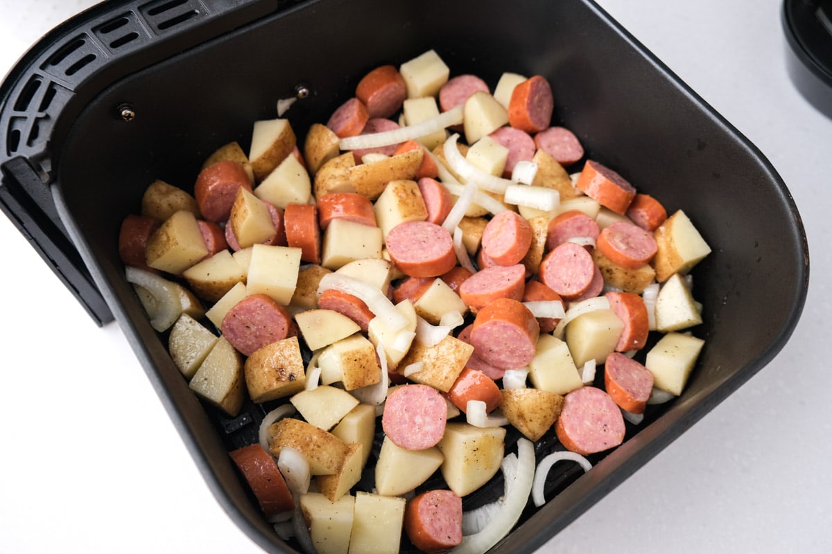 diced potatoes sausage pieces and onions in black air fryer tray sitting on white counter.