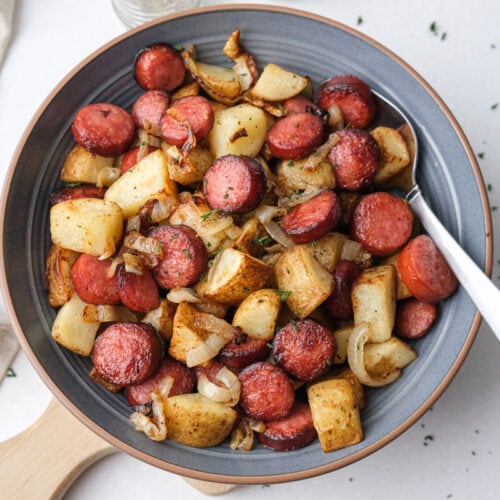 fried potatoes and sausage pieces in blue bowl with silver spoon on counter.