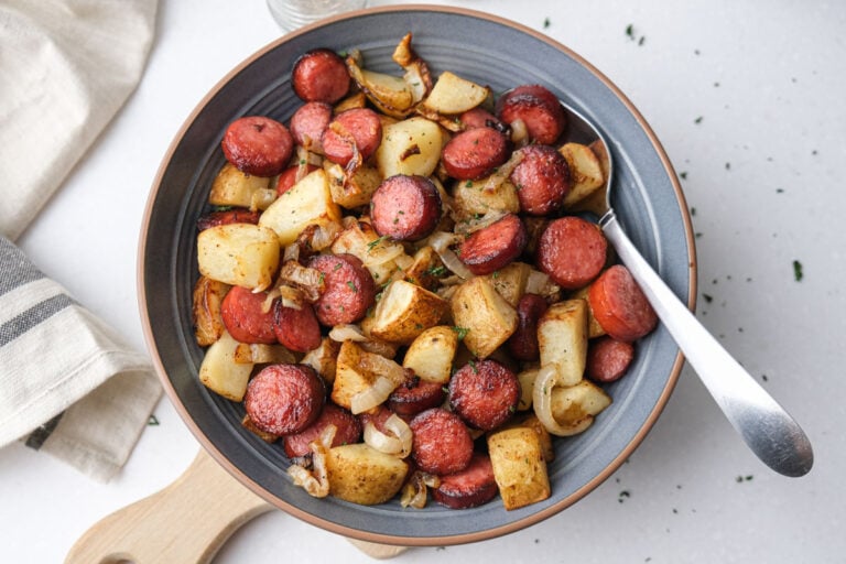 fried potatoes and sausage pieces in blue bowl with silver spoon on counter.