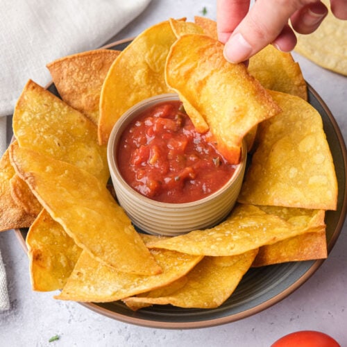 crispy tortilla chips around bowl of salsa with hand dipping one chip in.