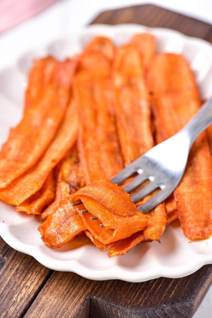 silver fork lifting strips of carrot bacon from white plate.