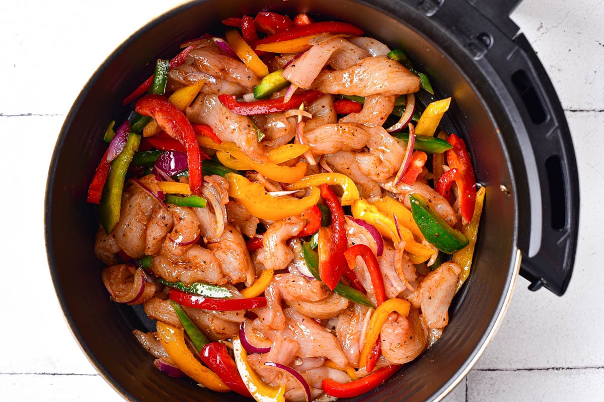 raw chicken peppers and onions covered in spices sitting in round black air fryer basket.