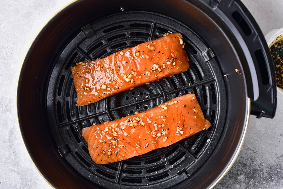two raw marinaded salmon fillets in round black air fryer basket.
