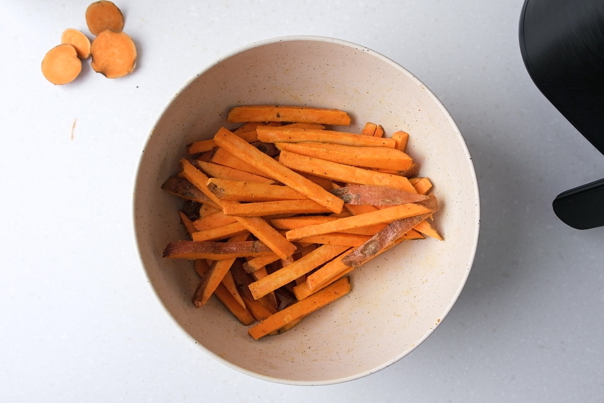 white mixing bowl of raw sweet potato fries with spices mixed in.