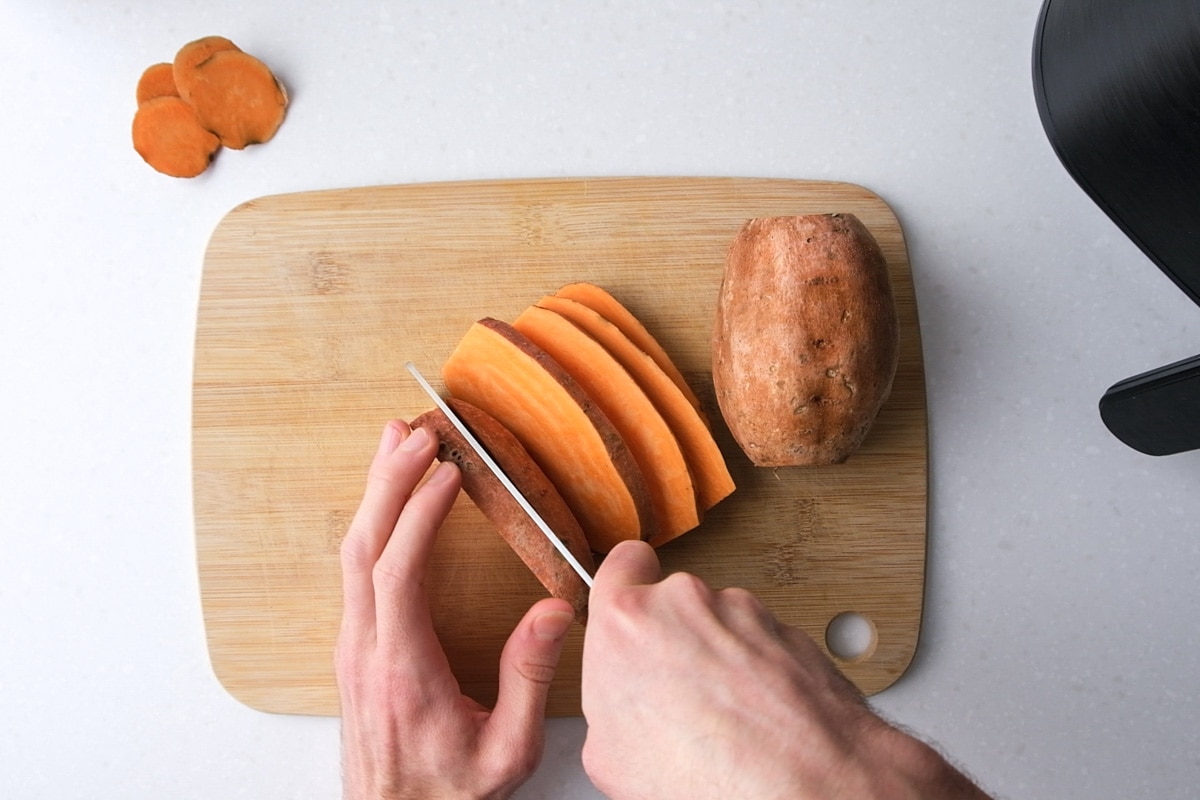 hand with knife cutting sweet potato into slices on wooden board.