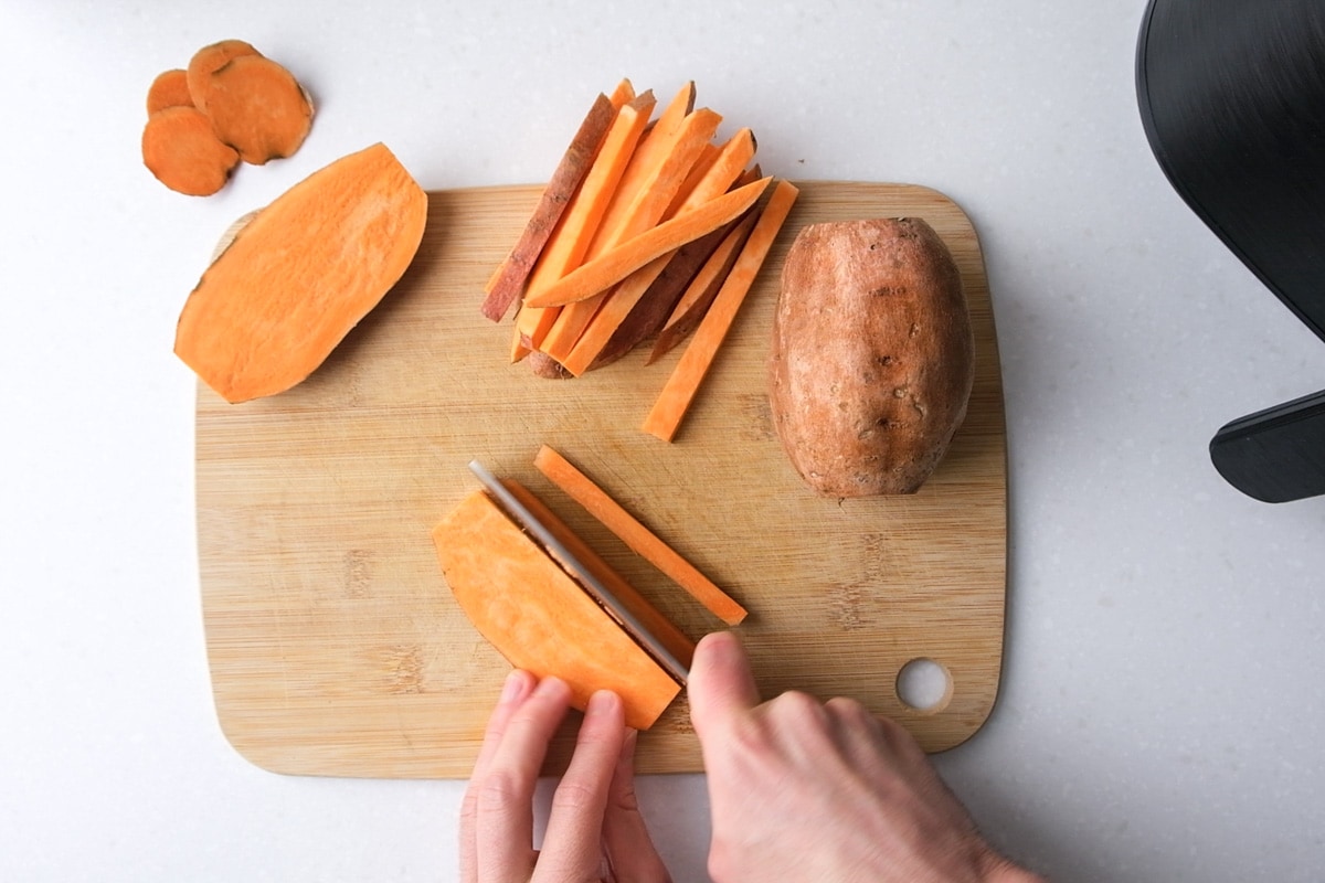 hand with knife cutting raw sweet potato into thin fries on wooden cutting board.