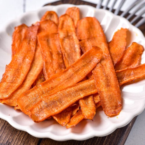 white plate of cooked carrot bacon on wooden board with forks beside.