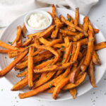 sweet potato fries on white plate with dip behind and chopped parsley around.
