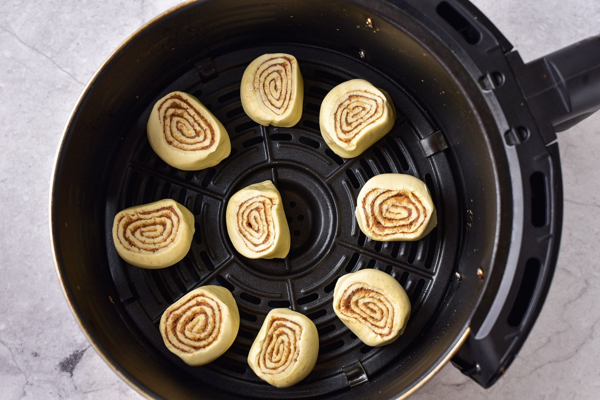raw cinnamon rolls arranged in a circle in round black air fryer tray on counter.