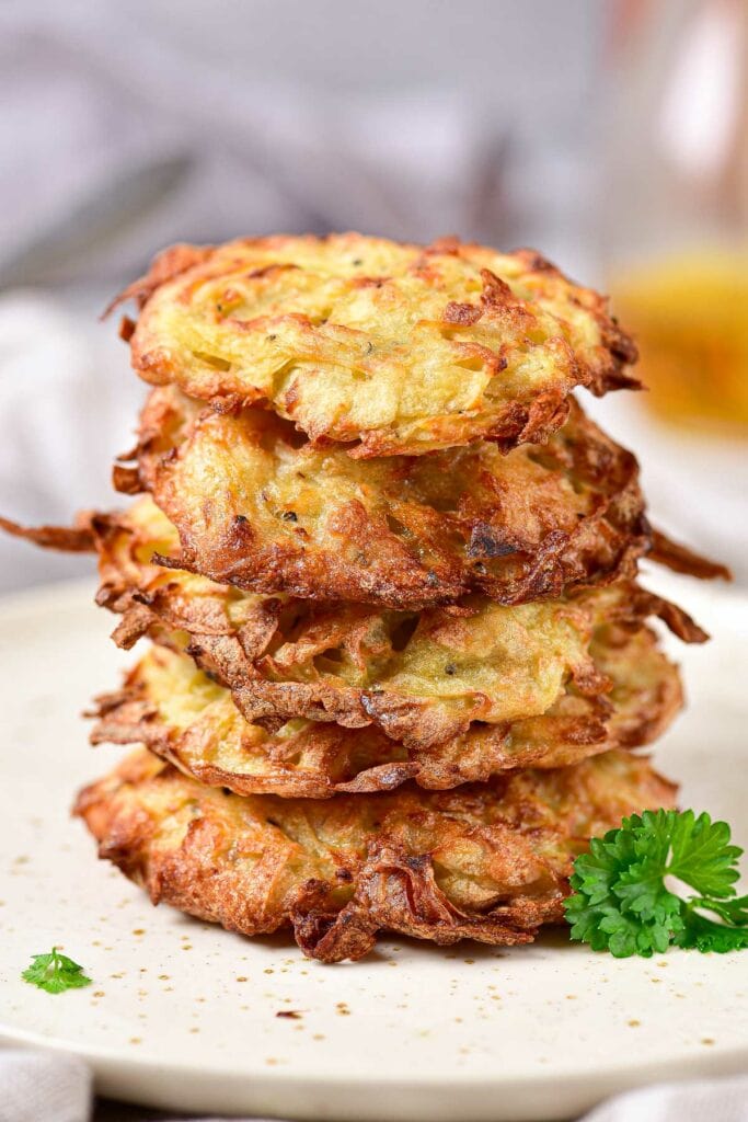 stack of crispy potato pancakes with green herbs beside on white plate.