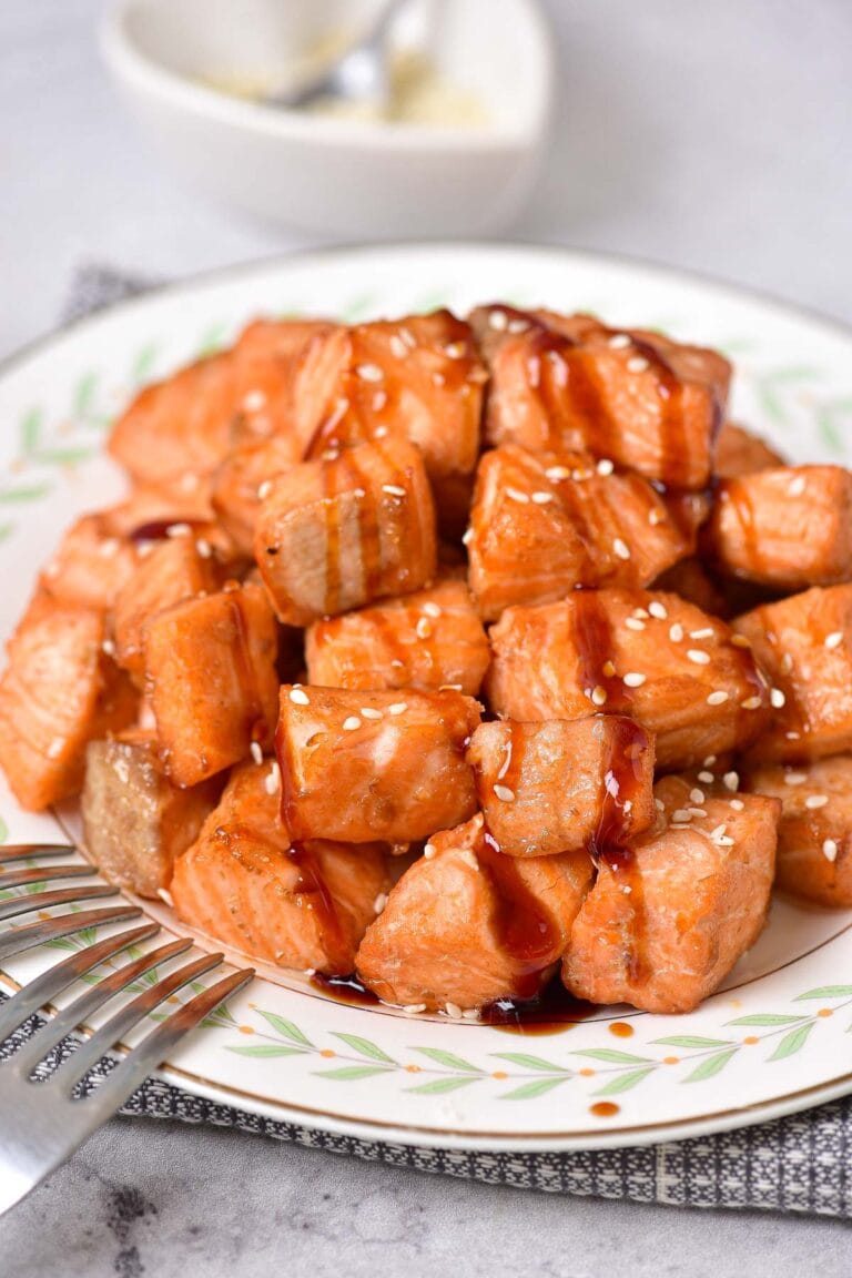 white plate with pile of salmon bites on it covered in drizzle and seeds with silver forks beside.
