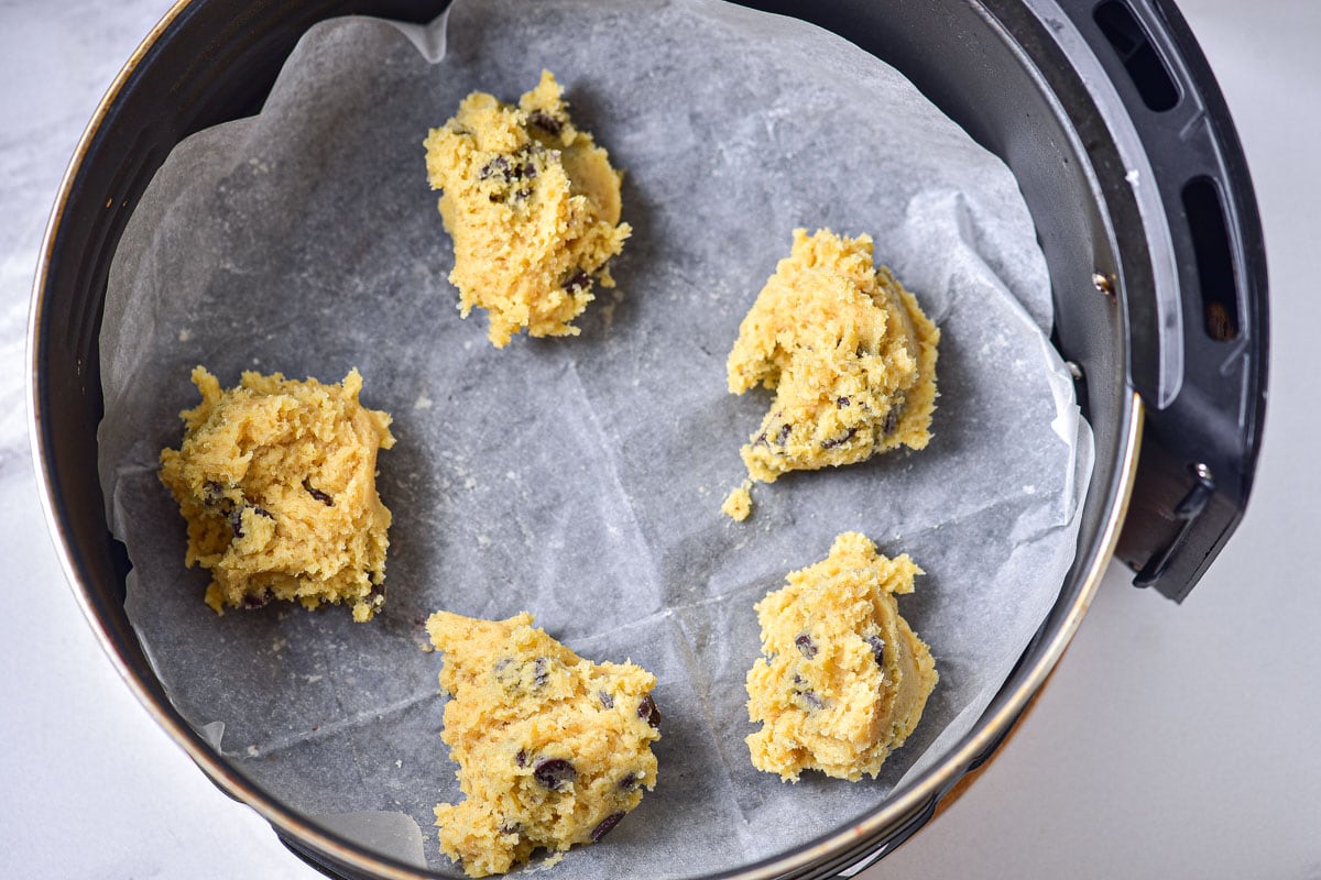 five lumps of raw cookie dough batter sitting on parchment paper in round air fryer basket.