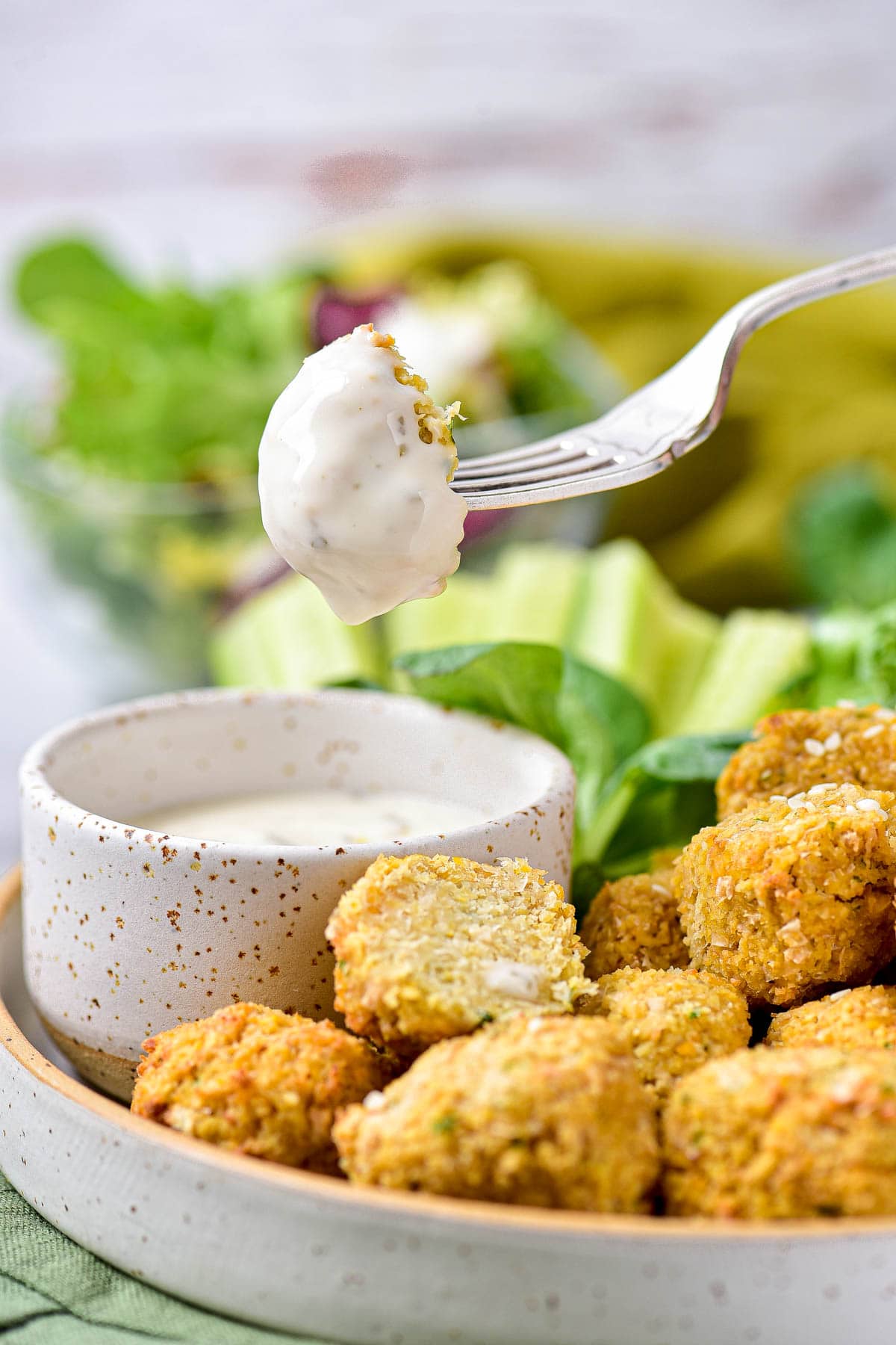 silver fork dipping half a falafel ball in white creamy dip with plate below.