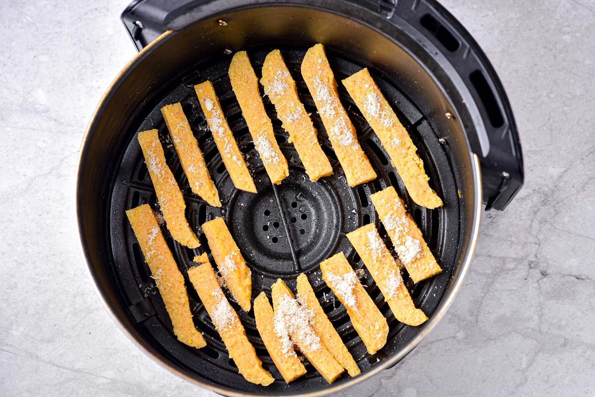 raw polenta fries in round air fryer basket with grated cheese on top.
